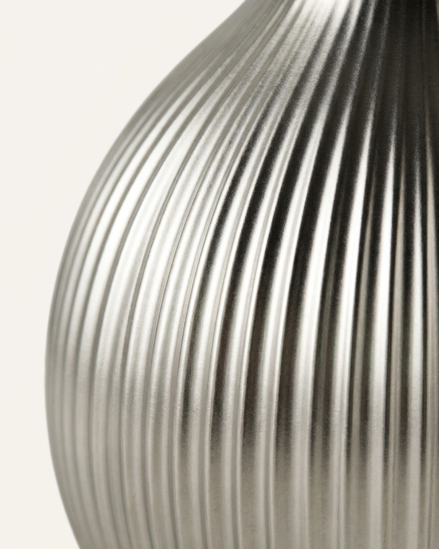 Close-up of a ribbed metallic silver object, likely a vase or decorative container, against a plain off-white background.