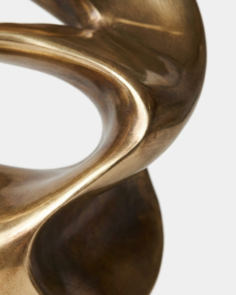 Close-up of a sculptural abstract object with a polished metallic bronze finish against a light gray background.