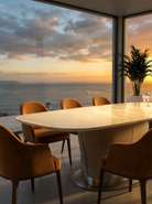 Modern dining area with marble table and chairs by large windows overlooking a sunset over water.