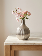 A ribbed metallic silver ceramic vase filled with pink roses and light pink blossoms, displayed on a light marble and wood side table against a warm beige wall.