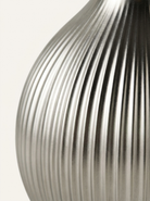 Close-up of a ribbed metallic silver object, likely a vase or decorative container, against a plain off-white background.