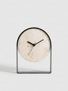 A modern minimalist tabletop clock with a circular beige stone dial and a sleek black metal frame, set against a clean light gray background.