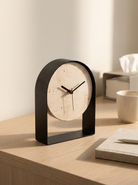 A modern arched tabletop clock with a black metal frame and a round beige stone-like dial, shown on a light wood desk beside a mug and notebook in a softly lit interior.