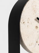 Close-up of a modern wall clock with a thick black frame and a circular natural travertine stone face on a light gray background.